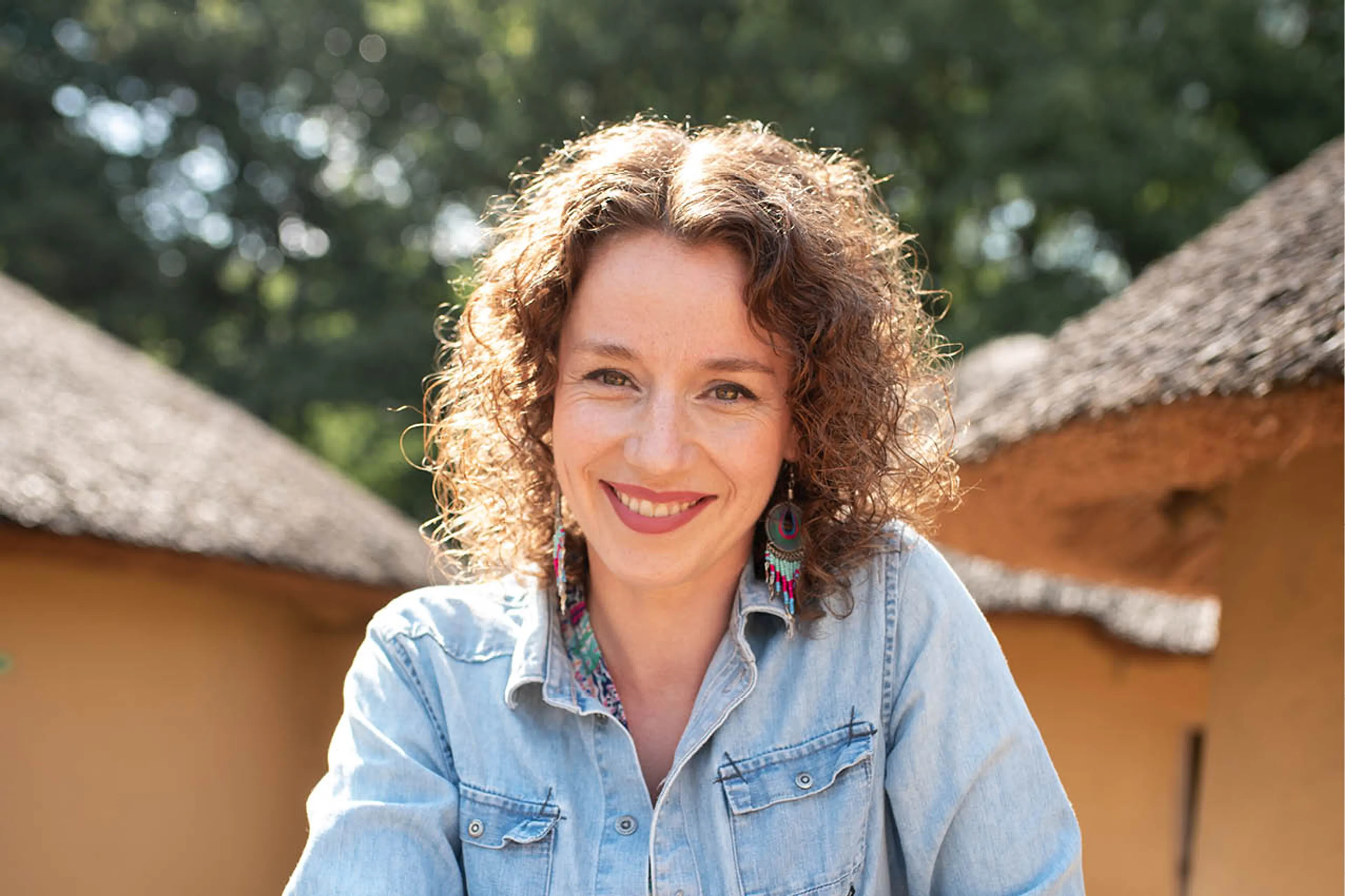 Annegina lacht in de camera. Ze heeft halflang krullerig bruin haar en een lichte spijkerblouse aan. Ze zit tussen twee huisjes in het Afrika Museum